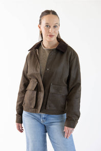A woman modeling a dark brown suede jacket with a dark brown corduroy collar, worn over a brown knit sweater and blue jeans, against a plain white studio background.