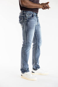 A person wearing light-wash blue denim jeans with decorative stitching and embroidered details on the back pockets, paired with white sneakers against a plain white studio background.