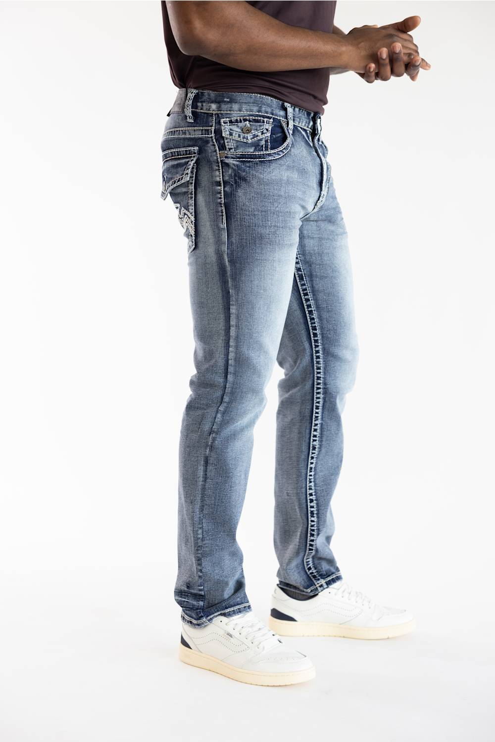 A person wearing light-wash blue denim jeans with decorative stitching and embroidered details on the back pockets, paired with white sneakers against a plain white studio background.