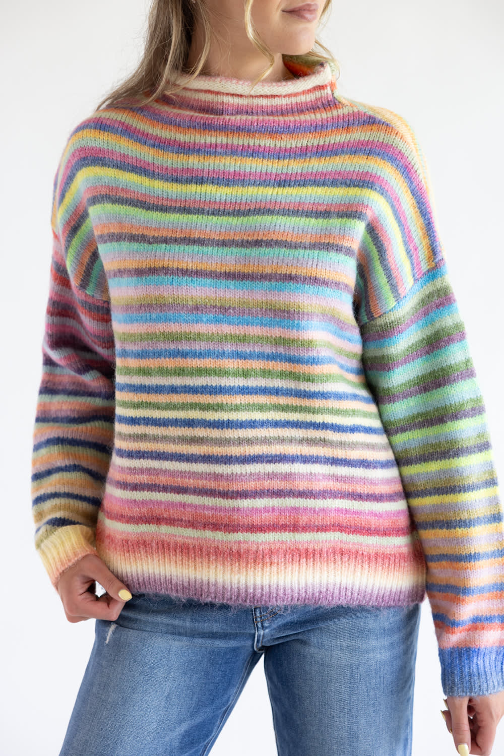 A close up of a rainbow striped multi colored long sleeve sweater with blue denim jeans standing in front of a white studio background
