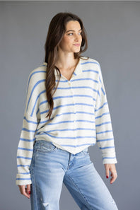 A woman modeling a blue and white striped knit button-up shirt with light-wash blue jeans, standing against a solid gray studio background.