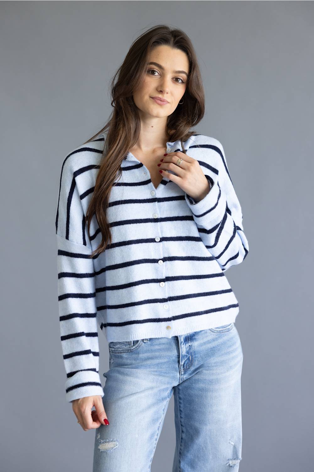 A woman modeling a white long-sleeve knit cardigan with navy blue horizontal stripes, paired with light-wash blue jeans, against a solid gray studio background.