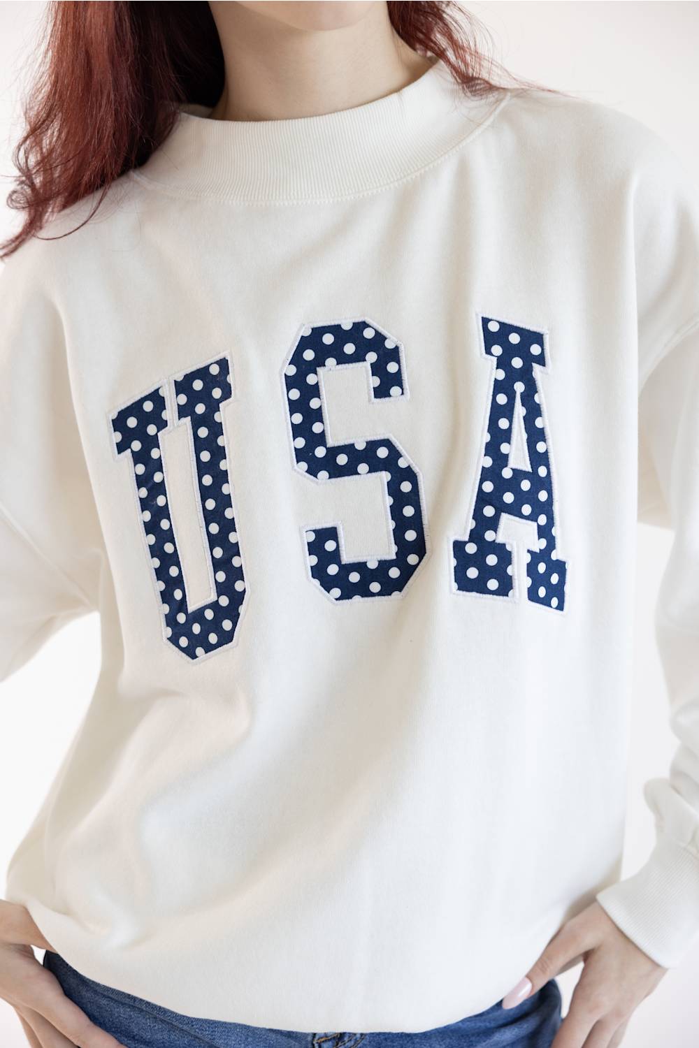A close-up of a person wearing a cream-colored crewneck sweatshirt with 'USA' printed across the chest in navy blue letters filled with white polka dots.