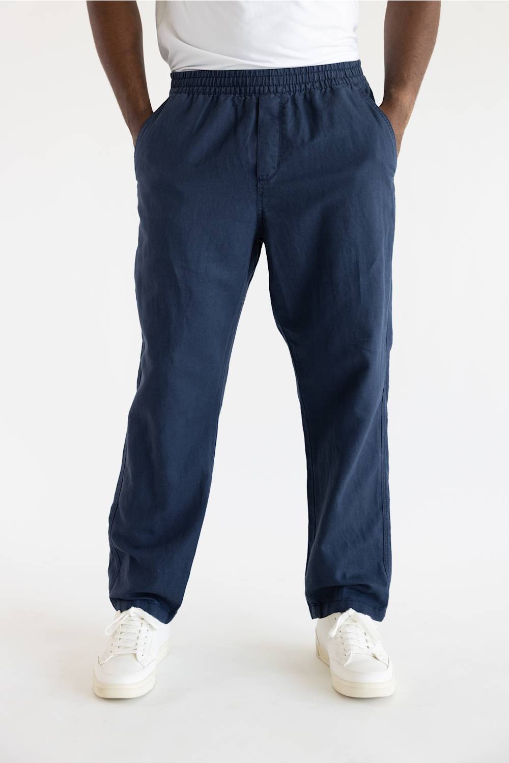 A person wearing dark navy blue jogger-style pants with an elastic waistband and side pockets, paired with a white t-shirt and white sneakers, shown from the waist down on a light gray studio background.