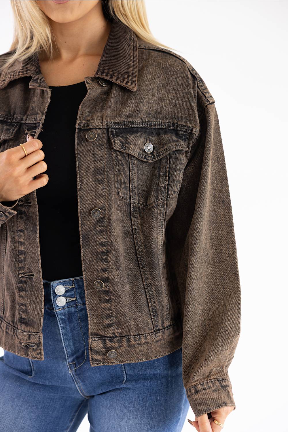 A woman wearing a washed dark brown denim jacket over a black top and blue jeans, shown from the neck down against a plain white background.