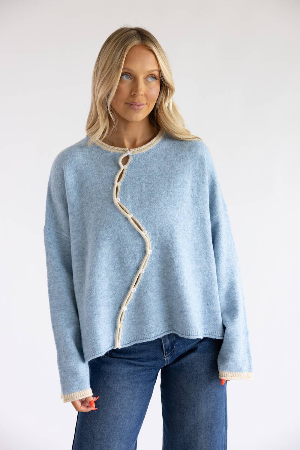 A woman wearing a light blue knit sweater with a distinctive wavy beige trim detail down the front, paired with dark blue jeans, against a plain white studio background.