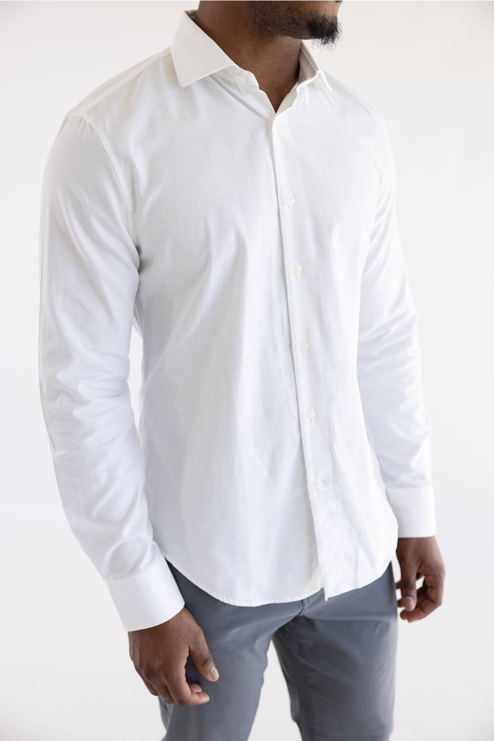 A man wearing a white long-sleeve button-down dress shirt with grey trousers, shown from the neck down against a plain white background.