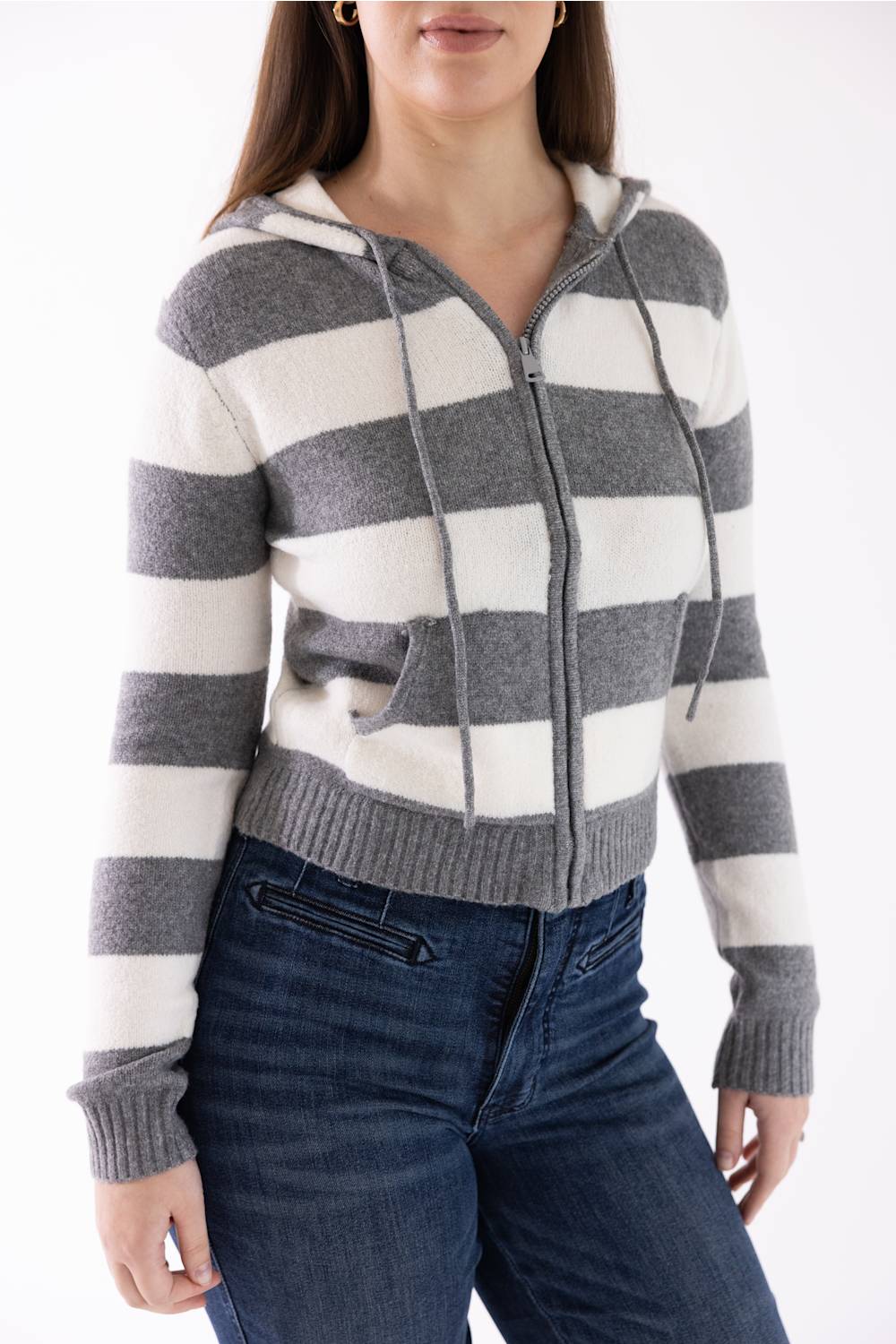 A woman wearing a cropped zip-up hoodie with horizontal gray and off-white stripes, paired with dark blue jeans, against a plain white background.