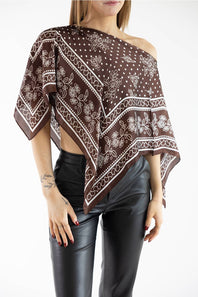 Woman wearing a brown and white patterned asymmetric scarf poncho top off one shoulder, styled with black leather pants against a plain white studio background.