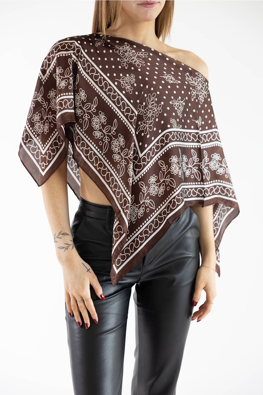 Woman wearing a brown and white patterned asymmetric scarf poncho top off one shoulder, styled with black leather pants against a plain white studio background.