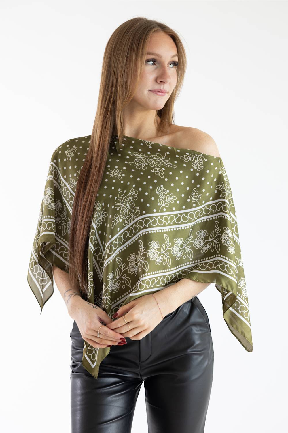 Medium shot of a woman modeling an off-the-shoulder olive green floral scarf poncho top with black leather pants.