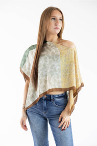 Woman modeling a green and yellow paisley patchwork off-the-shoulder scarf poncho top paired with denim jeans.