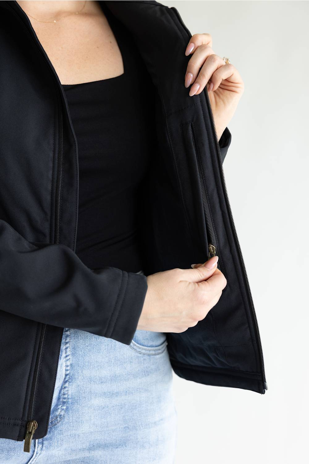 A close-up of a person wearing a black zip-front jacket over a black top and light blue jeans, opening the jacket to show the interior pocket area.