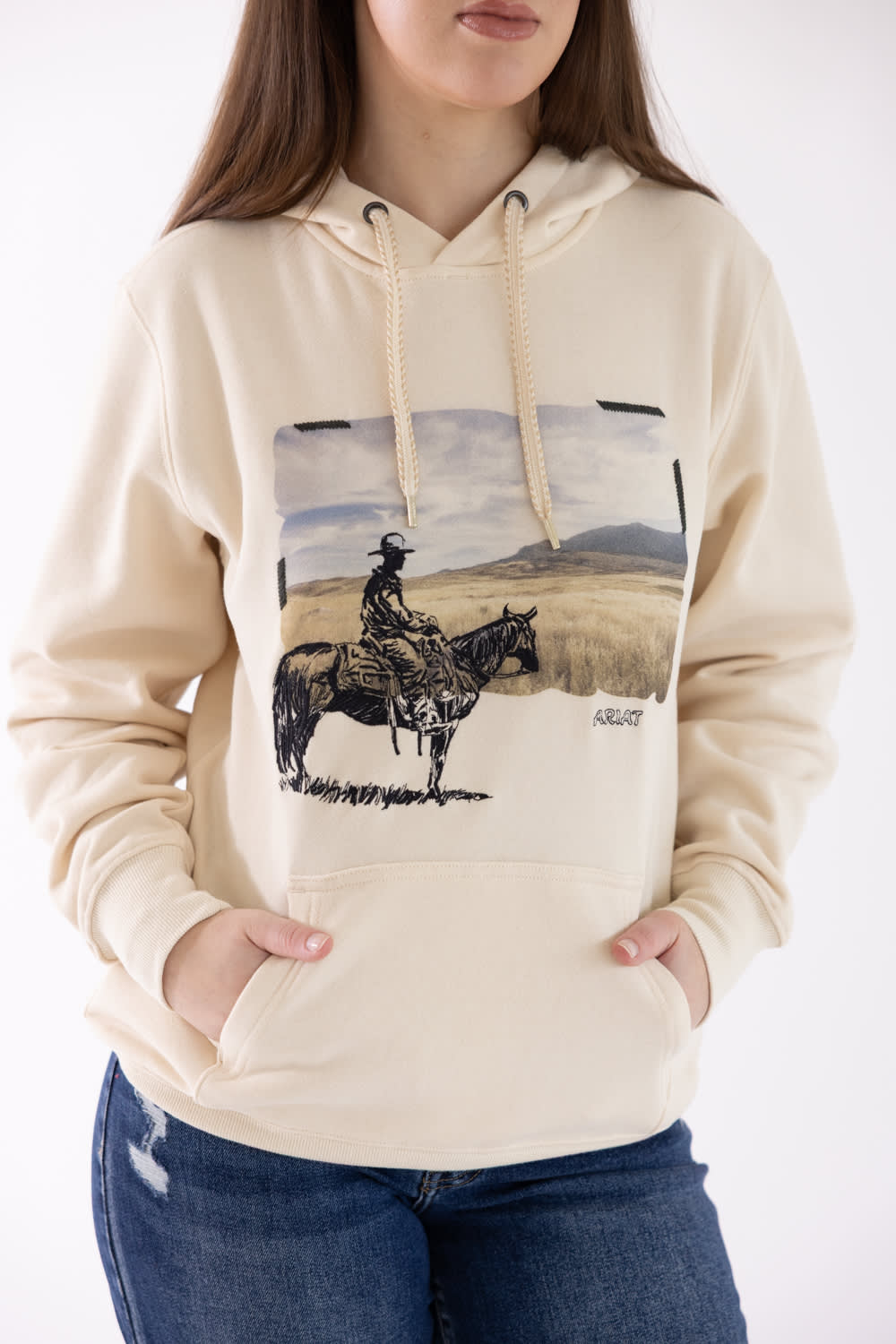 Front view of a model wearing the Ariat Wish You Were Here Hoodie in parchment beige, featuring a vintage western graphic of a cowboy on horseback, drawstrings, a kangaroo pocket, and rib-knit cuffs.