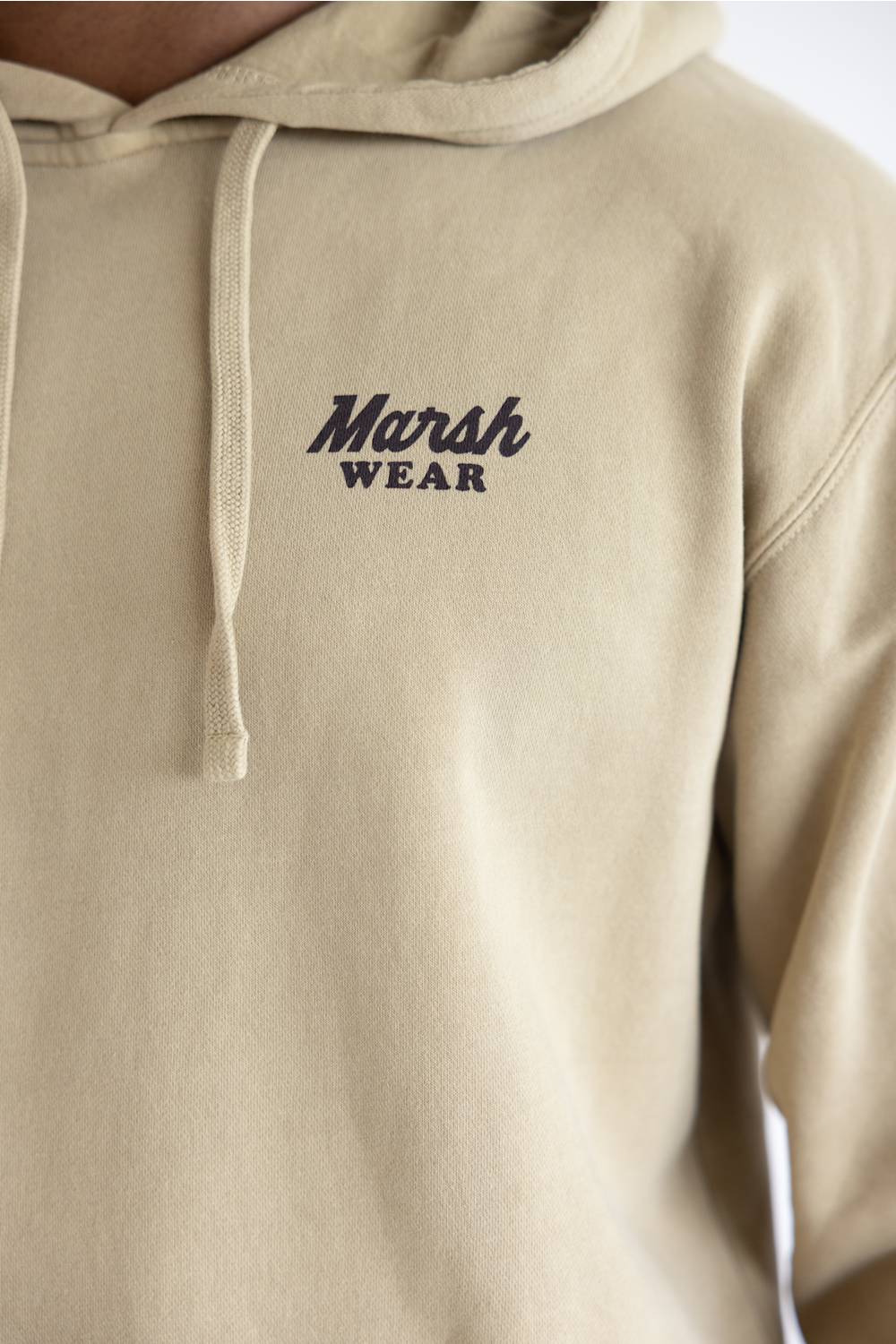 Close-up of a beige hoodie with the 'Marsh Wear' logo printed in dark navy on the left chest, featuring a drawstring hood and ribbed cuffs.