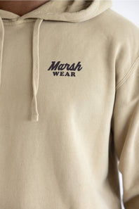 Close-up of a beige hoodie with the 'Marsh Wear' logo printed in dark navy on the left chest, featuring a drawstring hood and ribbed cuffs.