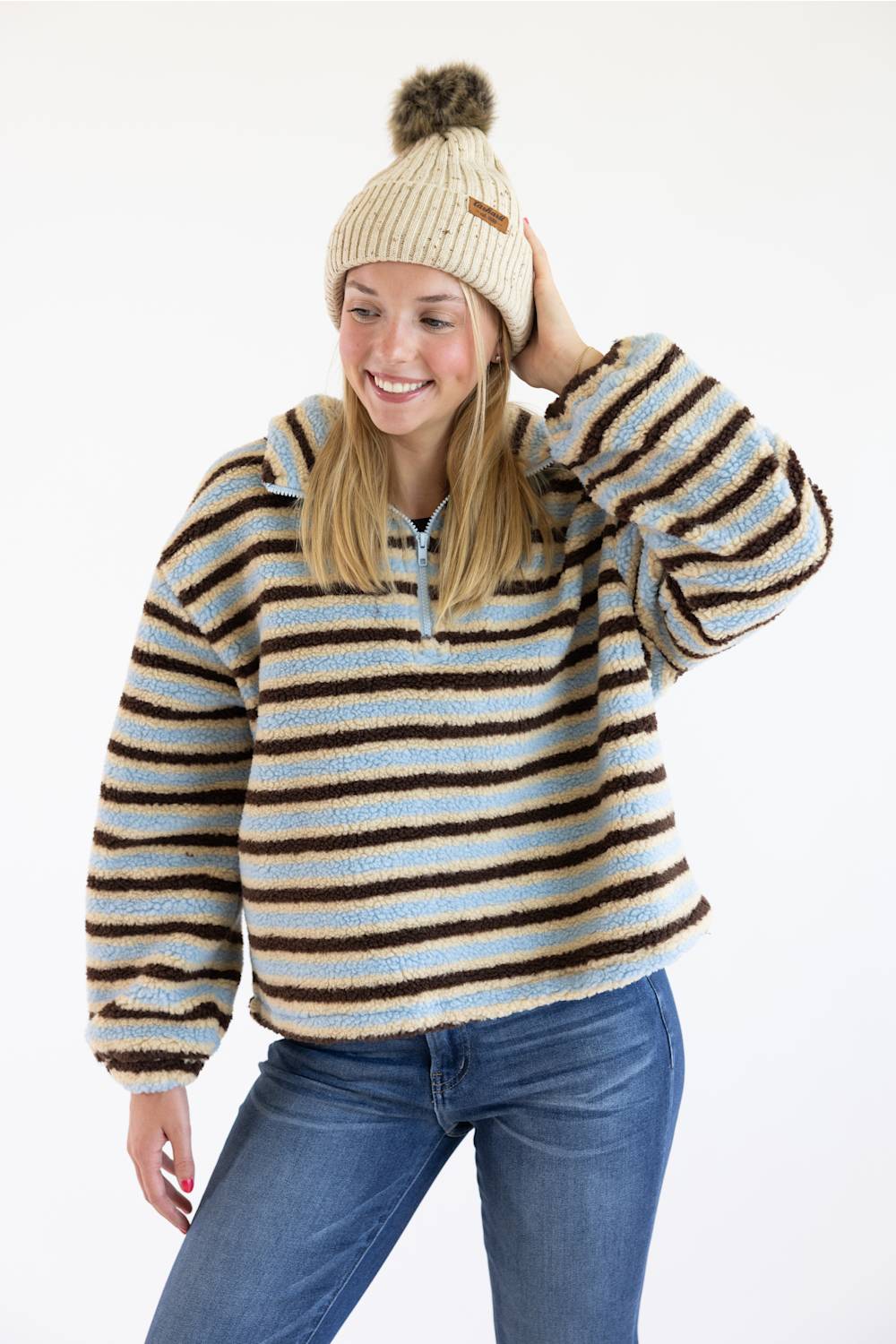 A woman wearing a fuzzy quarter-zip pullover sweater with horizontal brown, beige, and light blue stripes, paired with a cream knit beanie with a brown pom-pom and blue jeans, against a plain white studio background.
