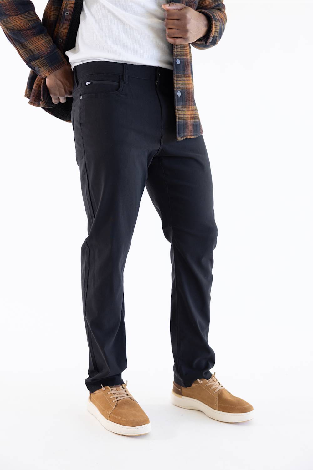 A person wearing slim-fit dark navy pants, styled with a white t-shirt, an open brown and navy plaid flannel shirt, and tan suede sneakers, shown from the waist down on a white studio background.