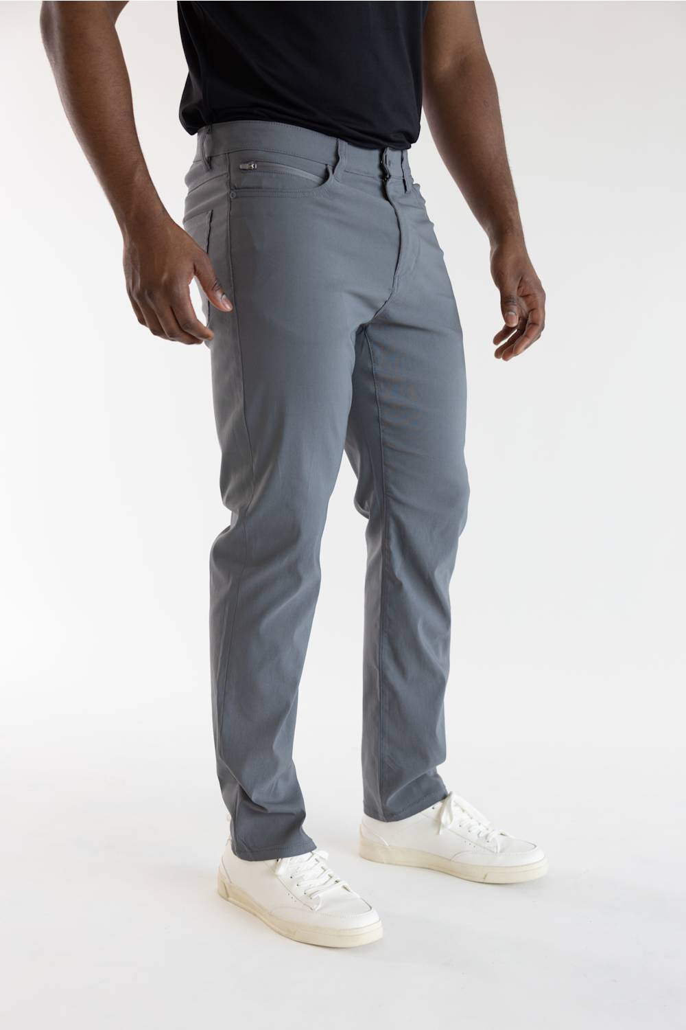 A man wearing slim-fit gray trousers, a black t-shirt, and white sneakers, shown from the waist down against a plain white studio background.