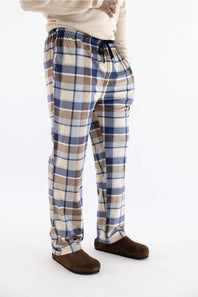 A person wearing plaid lounge pants in blue, beige, and white, paired with a cream sweater and brown slippers, shown from the waist down on a white studio background.
