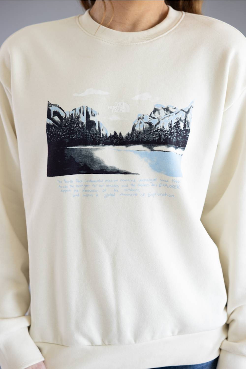 A person wearing a cream-colored crewneck sweatshirt with a graphic print of a mountain landscape and handwritten text on the front.