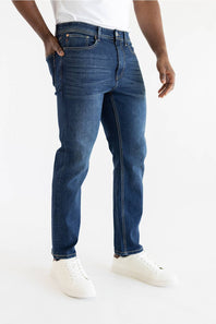 A person wearing dark blue slim-fit jeans with white sneakers, shown from the waist down against a clean white studio background.