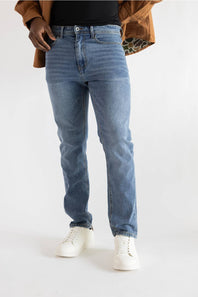 A person wearing light blue slim-fit jeans, styled with a black top, a brown jacket, and white sneakers, photographed against a plain white studio background.