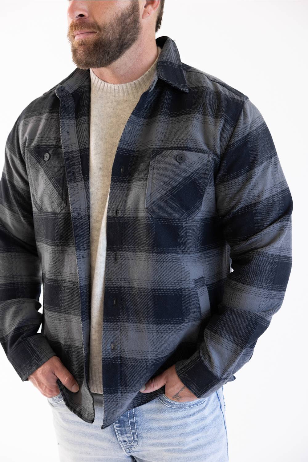 Close-up of a man wearing the Weatherproof Vintage Lumberjack Plaid Shirt Jacket in Phantom Night (charcoal grey and dark navy plaid), layered open over a beige ribbed knit sweater and light-wash jeans.