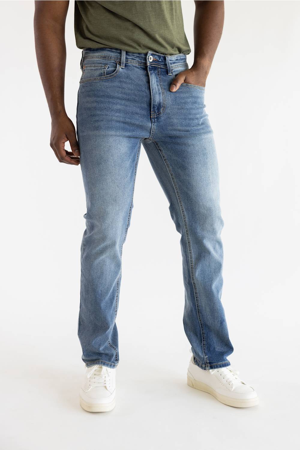 A person wearing light blue slim-fit denim jeans with a faded wash, paired with an olive green t-shirt and white sneakers, shown from the waist down on a white studio background.