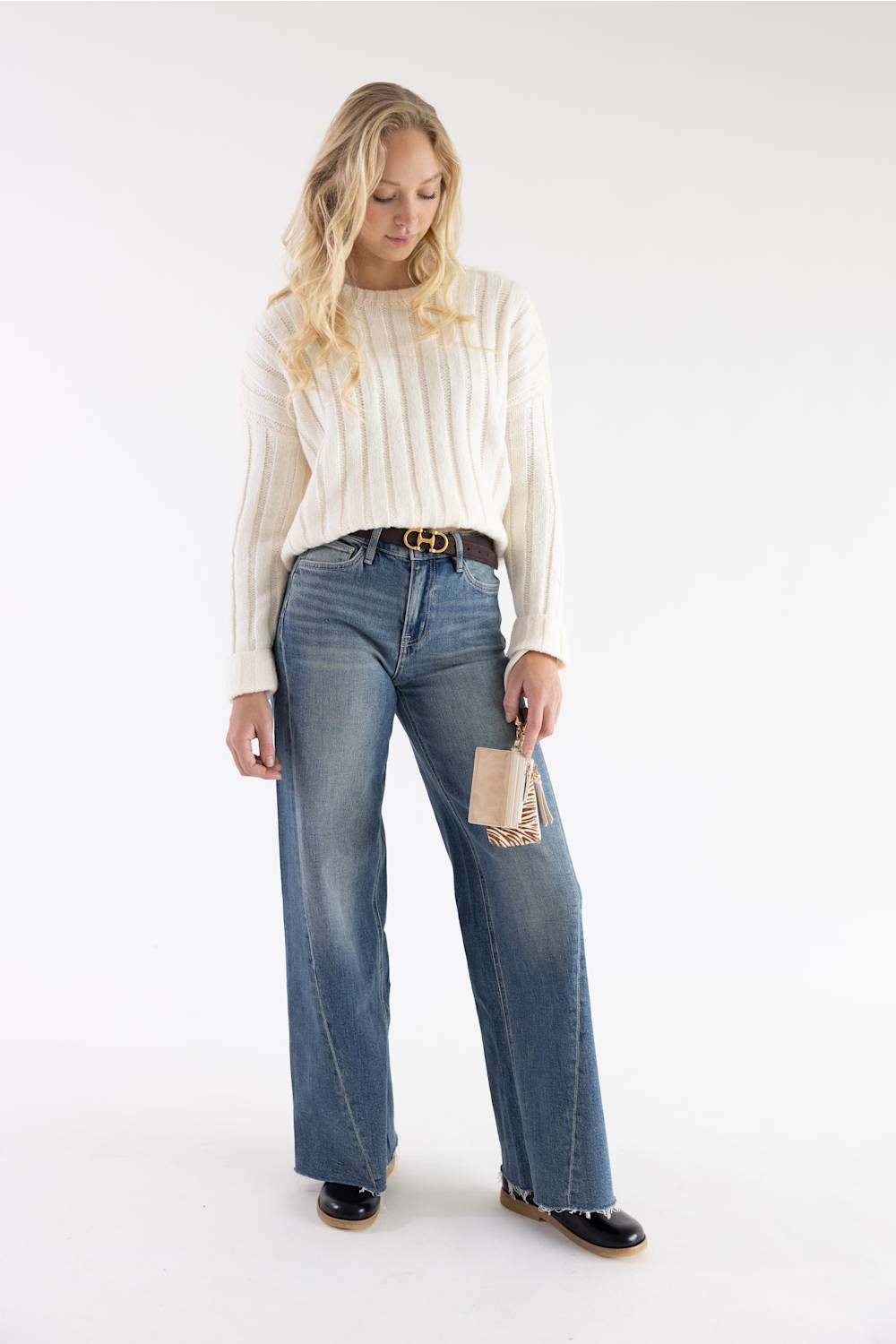 A woman modeling a cream ribbed knit sweater, blue wide-leg jeans, a brown belt, and black shoes against a plain white studio background.