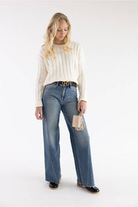 A woman modeling a cream ribbed knit sweater, blue wide-leg jeans, a brown belt, and black shoes against a plain white studio background.