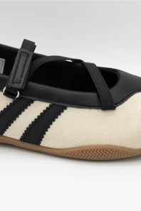 A close-up of a Mary Jane shoe featuring a cream suede upper, black leather trim and straps, three black stripes, and a brown gum sole.