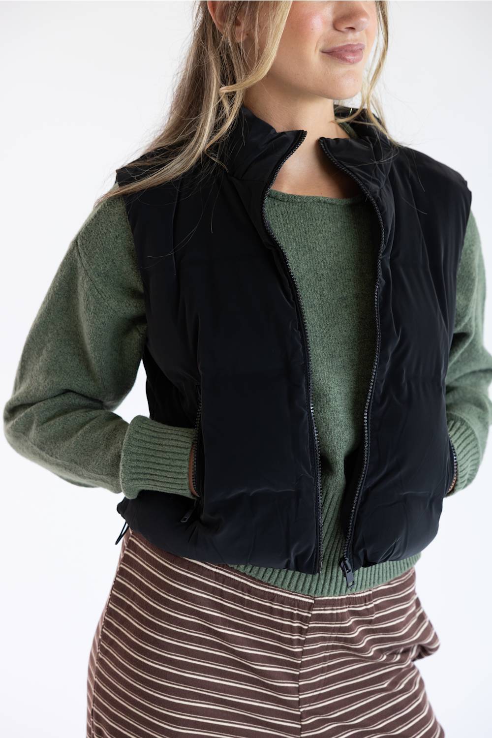 A woman wearing a black sleeveless zip-front puffer vest over an olive green knit sweater, paired with a brown and beige striped skirt, against a plain white background.