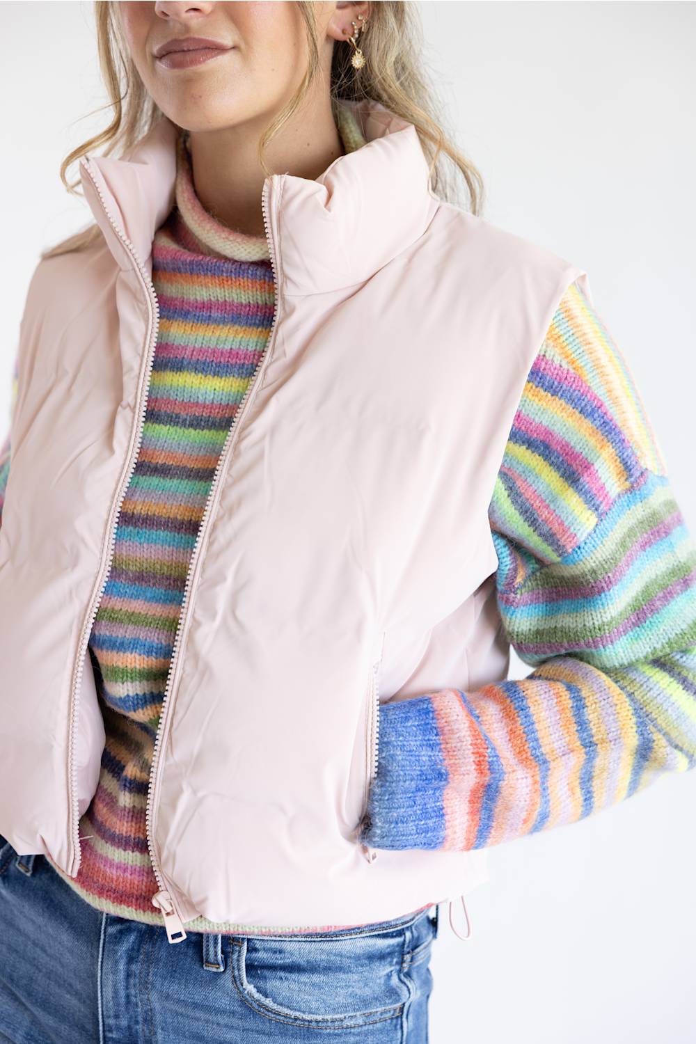 A woman wearing a pale blush pink puffer vest over a multicolored striped knit sweater and blue jeans, shown from the chest down against a plain light background.