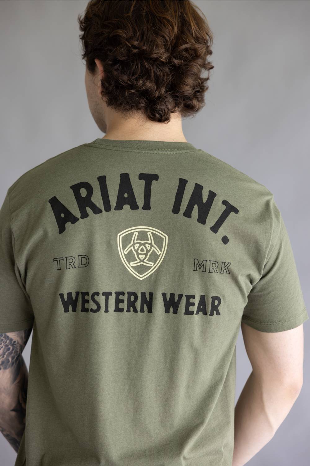 A man viewed from behind wearing an olive green t-shirt with a large black and beige graphic print on the back reading 'ARIAT INT. WESTERN WEAR' with a central shield logo.