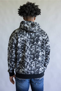 A man viewed from behind wearing a black and white abstract patterned hoodie with black ribbed cuffs and hem, paired with blue jeans, against a plain light gray background.