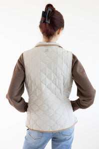 A woman shown from behind wearing a light beige quilted vest over a brown long-sleeve shirt and blue jeans, against a plain white background.