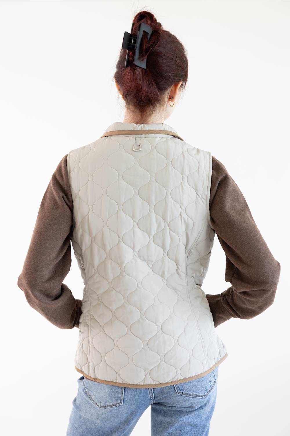 A woman shown from behind wearing a light beige quilted vest over a brown long-sleeve shirt and blue jeans, against a plain white background.