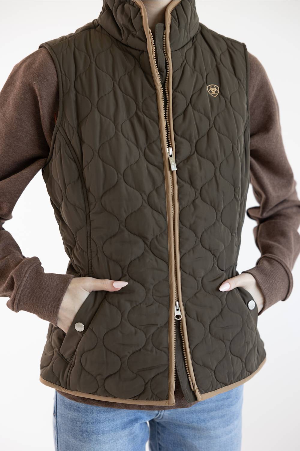 A woman wearing a dark brown quilted zip-front vest over a brown long-sleeve top and blue jeans, shown from the neck down against a light gray studio background.
