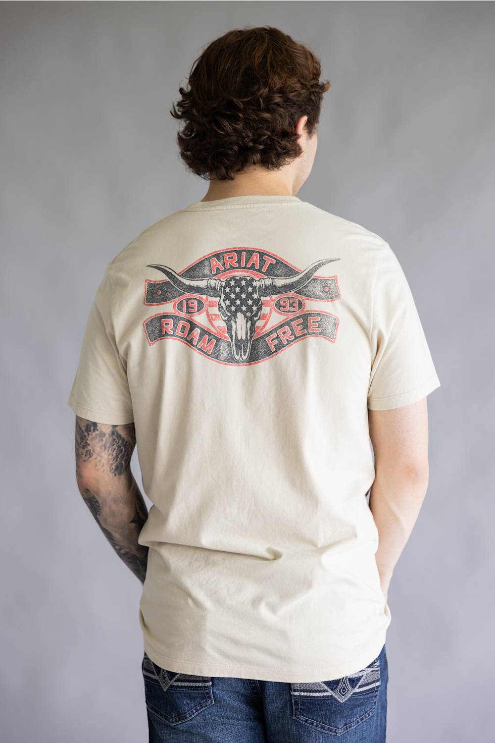 A man viewed from behind wearing a cream-colored t-shirt with a large graphic on the back featuring a steer skull and the text 'ARIAT ROMANCE FREE'.