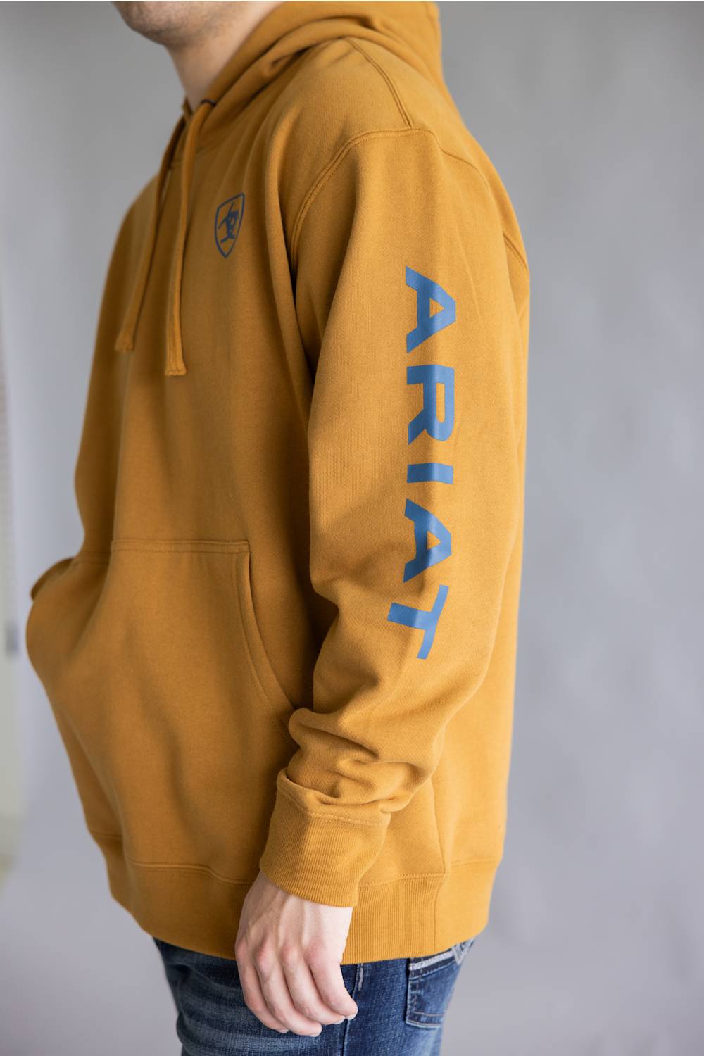 A person wearing a mustard yellow hoodie with a blue vertical 'ARIAT' logo on the left sleeve and a small blue shield logo on the left chest, paired with blue jeans against a gray background.