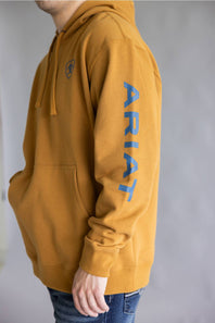 A person wearing a mustard yellow hoodie with a blue vertical 'ARIAT' logo on the left sleeve and a small blue shield logo on the left chest, paired with blue jeans against a gray background.