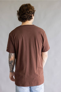 A man viewed from behind wearing a brown short-sleeve t-shirt and light blue jeans, standing against a plain gray studio background.