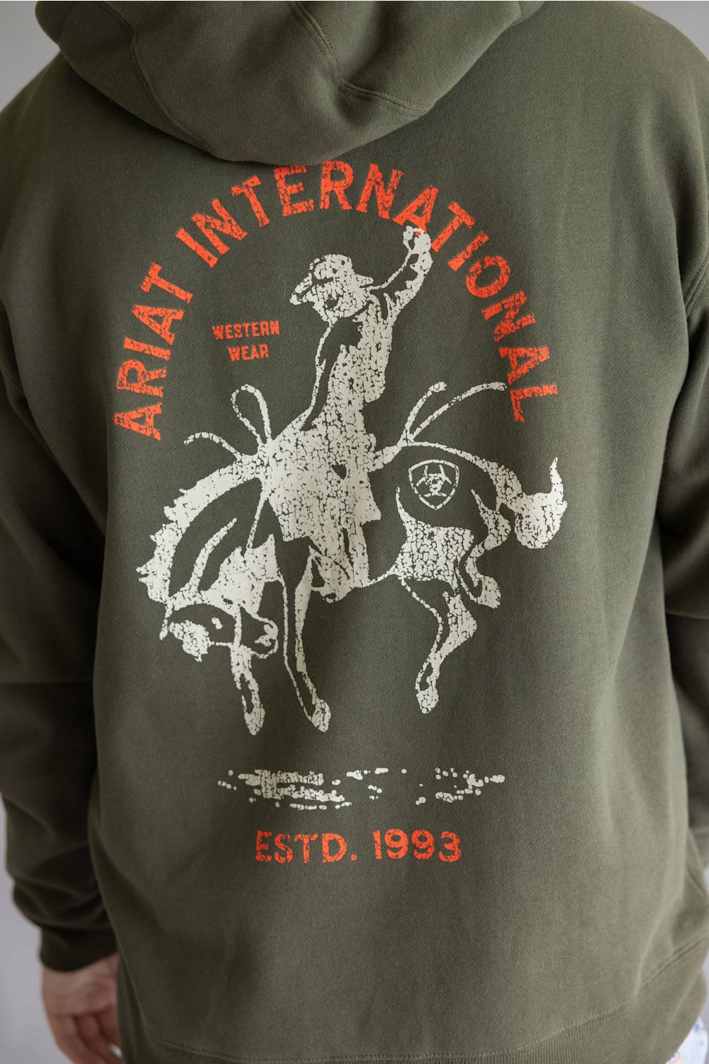 Close-up of the back of an olive green hoodie featuring a distressed white graphic of a cowboy riding a bucking bronco, with orange text reading 'ARIAT INTERNATIONAL' and 'ESTD. 1993'.