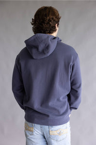 A person viewed from behind wearing a dark slate blue hoodie and light blue jeans with decorative stitching on the back pockets, set against a plain gray studio background.