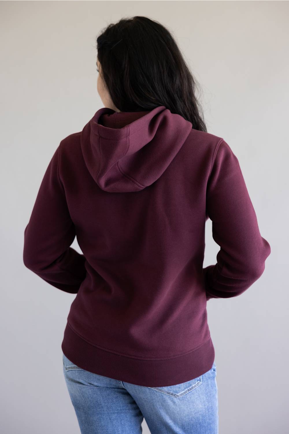 A woman viewed from behind wearing a maroon hooded sweatshirt and light blue jeans against a plain light gray wall.