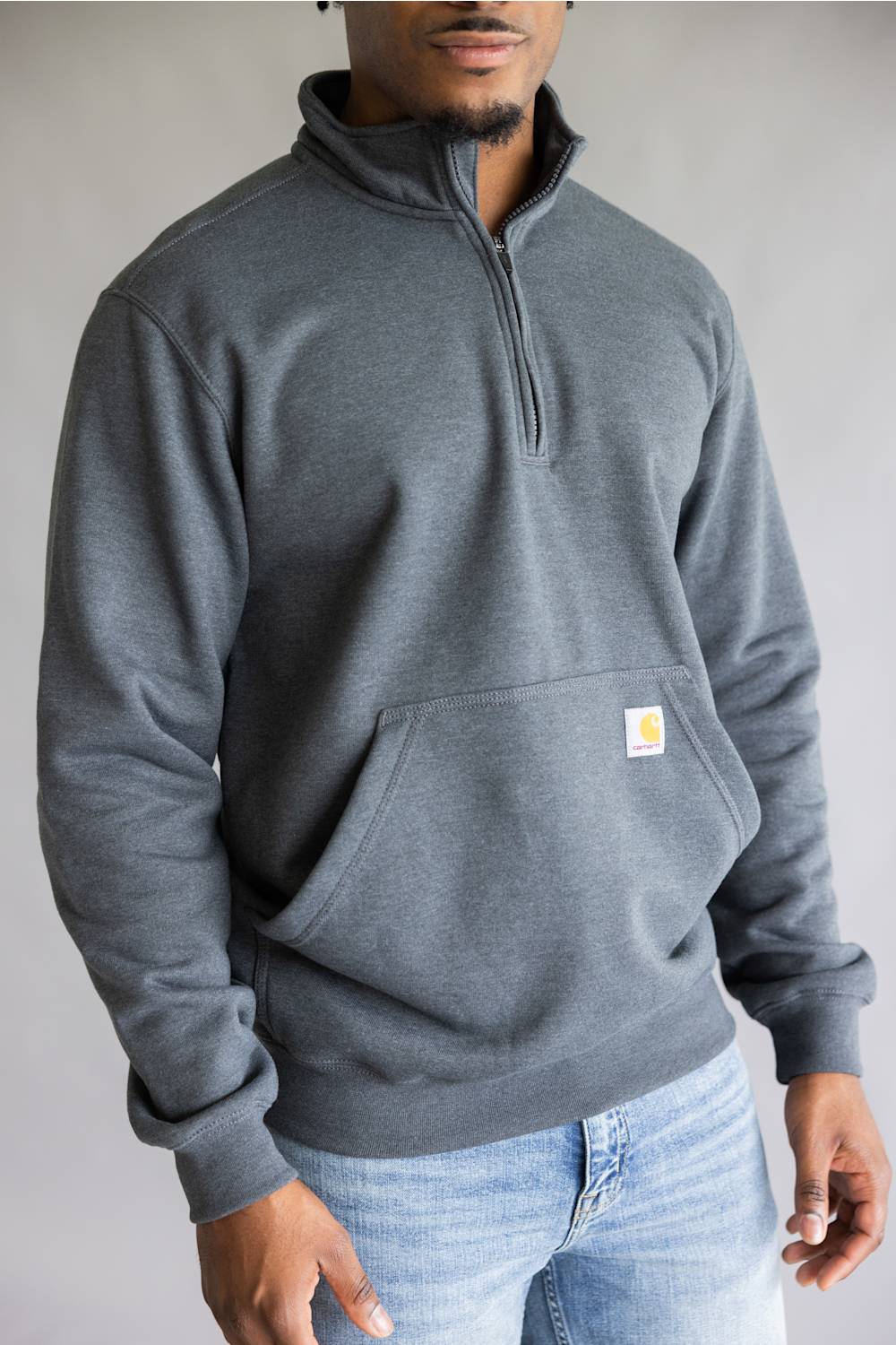 A man wearing a dark gray heathered quarter-zip sweatshirt with a small Carhartt logo patch on the chest, paired with light blue jeans, against a neutral gray background.