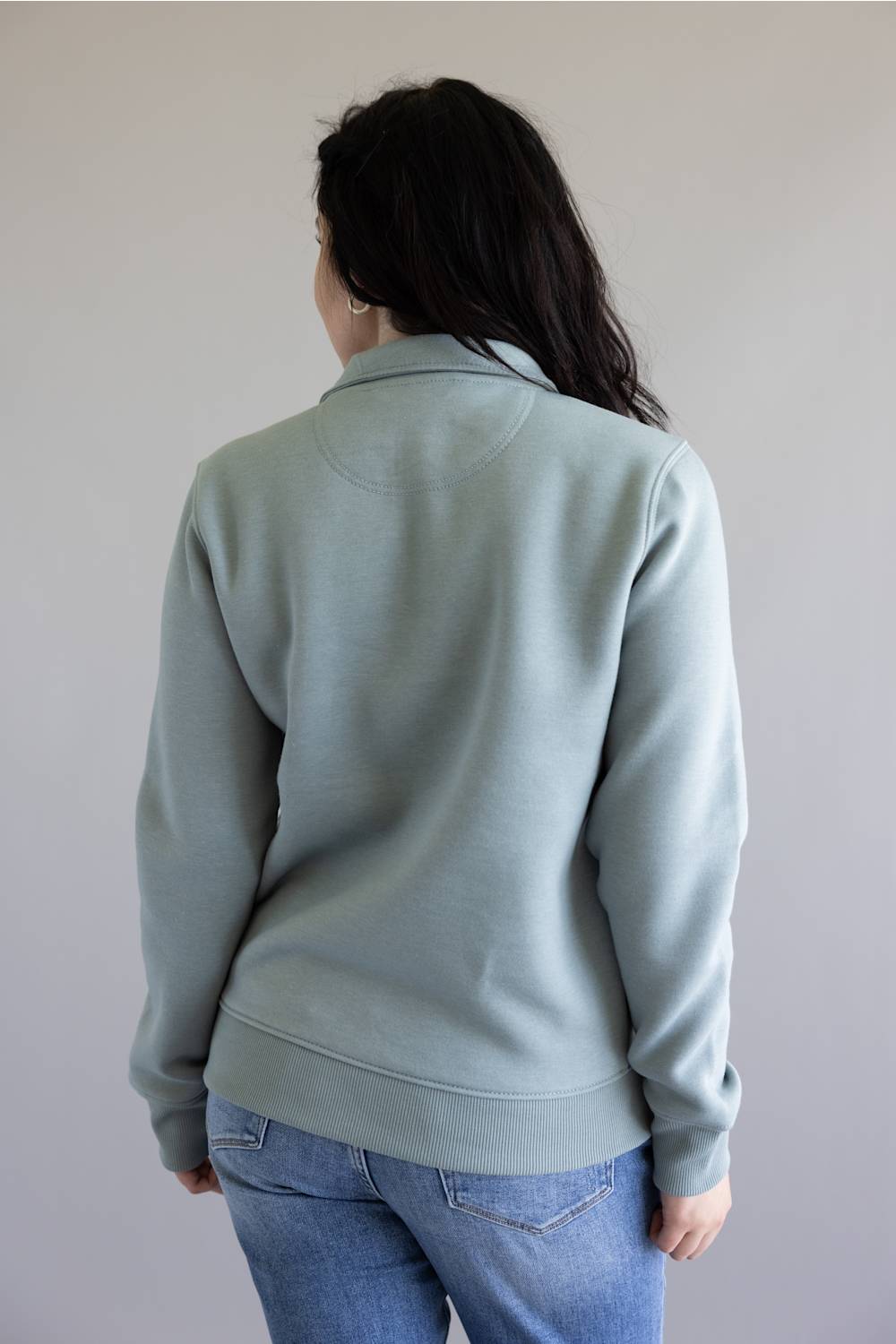 A woman viewed from behind wearing a pale sage green long-sleeve sweatshirt and blue jeans against a plain light gray wall.