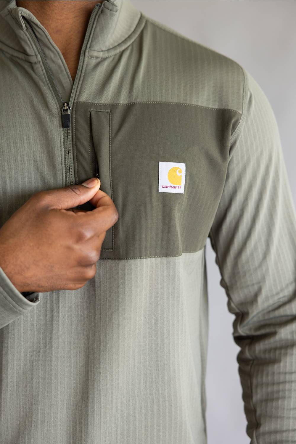 Close-up of a person wearing a quarter-zip pullover in a muted sage green color, featuring a contrasting darker green chest pocket with a visible Carhartt logo patch.