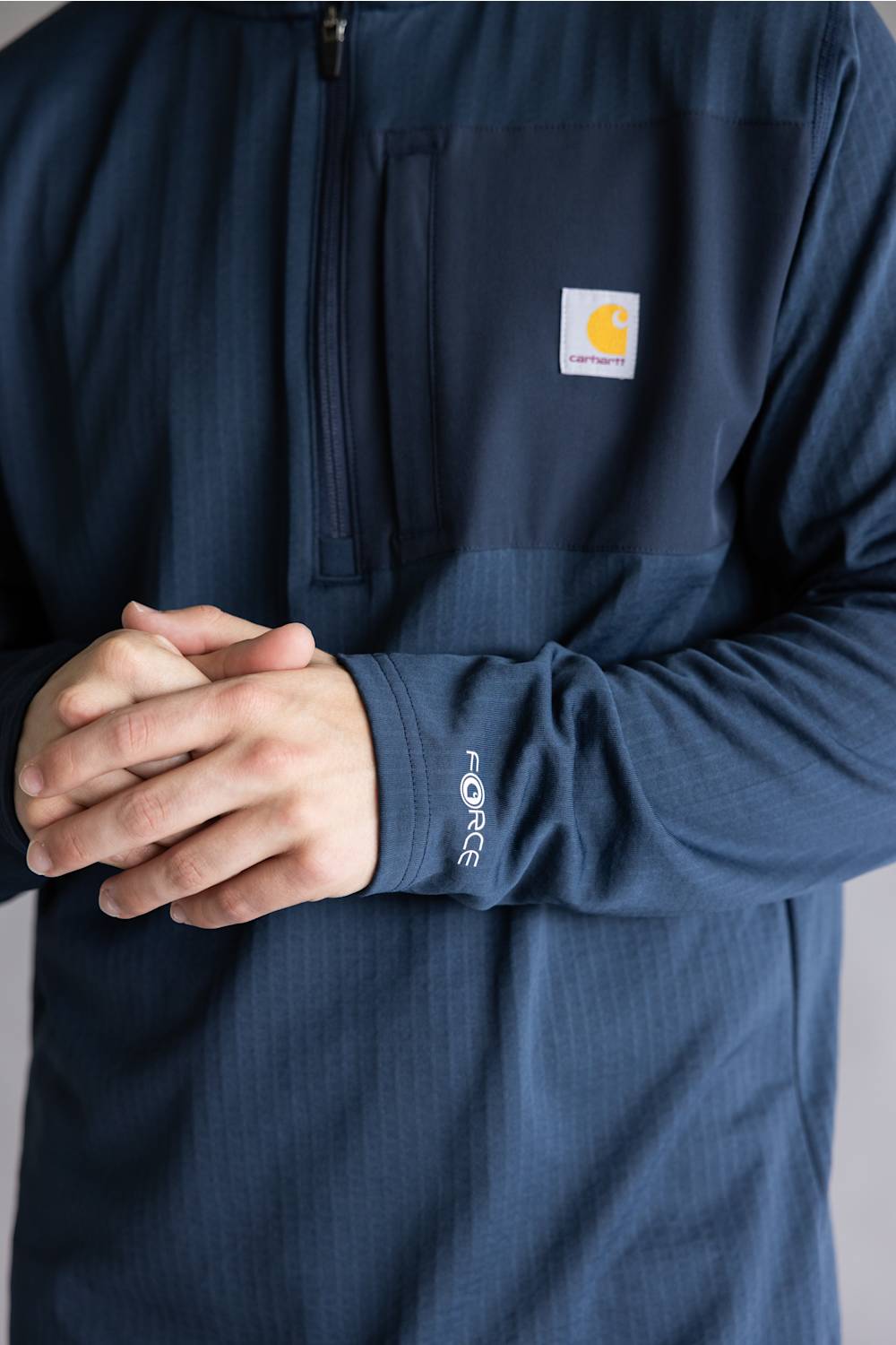 A close-up of a person wearing a navy blue long-sleeve quarter-zip pullover with a chest pocket and a visible Carhartt logo patch. The person's hands are clasped in front of the torso.
